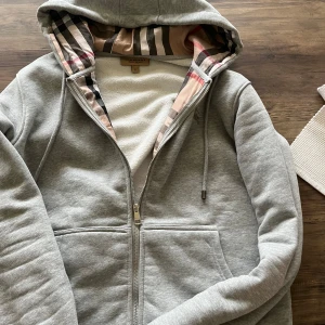 Grå hoodie från Burberry - Säljer en stilren grå hoodie från Burberry med dragkedja och klassiskt rutigt mönster i huvan. Tröjan har en broderad logga på bröstet och är tillverkad i mjuk bomull. Perfekt för en avslappnad look. 