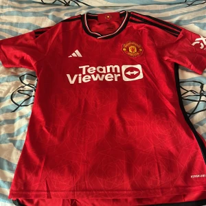Röd Manchester United fotbollströja från Adidas - Säljer en röd Manchester United fotbollströja från Adidas med korta ärmar och svartvita detaljer. Tröjan var en gåva, men jag bär inte på storleken L vilket gör att tröjan ser för stor ut på mig. Tänkte om det var någon som kan använda det mer än vad jag någonsin har gjort ❤️😊