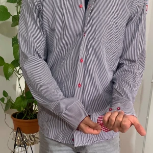 Randig skjorta från Oscar of Sweden - Snygg randig skjorta från Oscar of Sweden i blått och vitt med röda knappar. Kragen har ett rödvitt rutigt mönster på insidan som ger en cool kontrast. Perfekt för en stilren look!