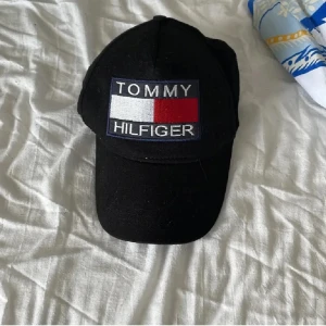 Svart keps från Tommy Hilfiger - Svart keps med böjd skärm och stort broderat Tommy Hilfiger-märke framtill i vitt, rött och blått. Justerbar baktill för bra passform. Perfekt accessoar för en sportig och stilren look.