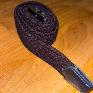 Mörkbrunt flätat bälte - Säljer ett mörkblått flätat bälte med detaljer i skinn och ett klassiskt spänne i metall. Bältet har en stilren design och passar perfekt till jeans eller chinos. Köptes i butik från friendhs för 400kr.