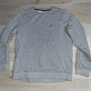 Grå sweatshirt från Lacoste - Säljer en klassisk grå sweatshirt från Lacoste med rund halsringning och den ikoniska gröna krokodilloggan broderad på bröstet. Tröjan har långa ärmar och ribbade muddar vid ärmslut och nederkant. Perfekt för en stilren och avslappnad look. Storleken är S eller 176 cm. Skriv gärna vid minsta fråga, pris kan diskuteras. Nypris runt 1100kr.
