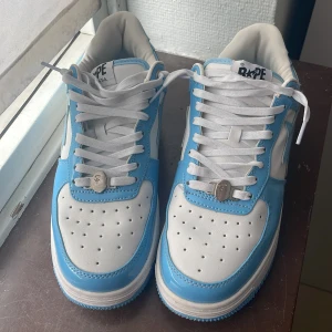 BAPE STA sneakers i blått och vitt - Säljer ett par BAPE STA sneakers i blått och vitt med klassisk stjärndesign på sidan. Skorna har snörning, rund tå och platt sula. Ovandelen är i blank syntet med vita detaljer och blå kontraster. Perfekta för dig som gillar streetwear och vill sticka ut.