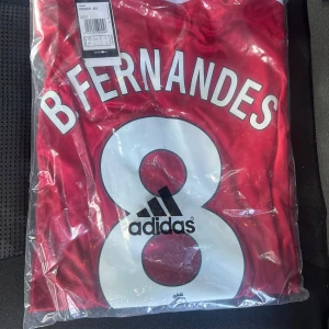 Manchester United fotbollströja B Fernandes 8 - Röd Manchester United fotbollströja med B Fernandes och nummer 8 på ryggen. Adidas-logga tryckt på siffran. Klassisk design med vita detaljer och rund hals. Perfekt för fans som vill visa sitt stöd på matchdagar.