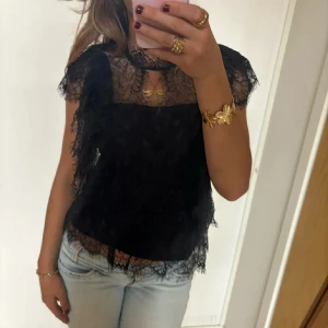 Svart spetsblus med volangdetaljer - Säljer en svart blus i spets med volangdetaljer och transparenta partier upptill. Blusen har korta ärmar och knäppning i nacken. Perfekt för dig som vill ha en feminin och trendig topp.
