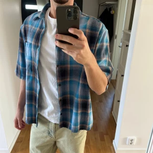 Blå och beige rutig kortärmad skjorta  - Säljer en blå och beige rutig skjorta från asos/collusion. Skjortan har en avslappnad passform och bröstficka. Perfekt för varmare dagar och enkel att styla över en t-shirt.