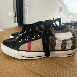 Burberry sneakers med klassiskt rutmönster - Säljer ett par Burberry sneakers med svart läderdetalj, klassiskt beige och svart rutmönster samt röda ränder. dålig innersula. säljer därför för billigt pris. Inte säker om äkta