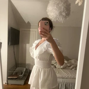 Vit volangklänning från Gina Tricot - Supersöt vit klänning från Gina Tricot i storlek XS. Klänningen har v-ringning, volangdetaljer vid axlar och kjol samt korta puffärmar. Tillverkad i ett lätt, strukturerat material som ger en luftig känsla. Perfekt för sommarens alla tillfällen!