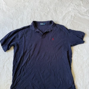 Mörkblå pikétröja från Polo Ralph Lauren - Säljer en klassisk mörkblå pikétröja från Polo Ralph Lauren med röd broderad logga på bröstet. Tröjan har korta ärmar, krage och knappar vid halsen. Perfekt för en stilren och avslappnad look. Finns ett hål på framsidan plus lite släpp i en söm. 