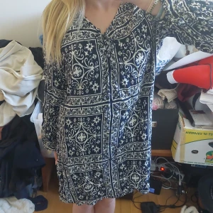 Mönstrad blåvit klänning med lång ärm - Säljer en blåvit klänning med lång ärm och vackert, detaljerat mönster. Klänningen har en rak passform och knäppning framtill. Perfekt för dig som gillar bohemiska och unika plagg.