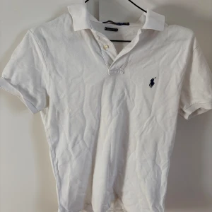 Vit pikétröja från Polo Ralph Lauren - Klassisk vit pikétröja från Polo Ralph Lauren med korta ärmar och broderad marinblå logga på bröstet. Tröjan har krage och två knappar vid halsen. Perfekt för en stilren och sportig look.