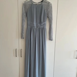 Ljusblå långklänning med spetsärmar och öppen rygg - Säljer en elegant ljusblå långklänning med plisserad överdel, spetsärmar och öppen rygg. Klänningen har ett brett midjeband med knyt bak och långa ärmar i spets. Perfekt för bal eller festliga tillfällen. Från Tfnc använd 1 gång på balen😁 Så snygg