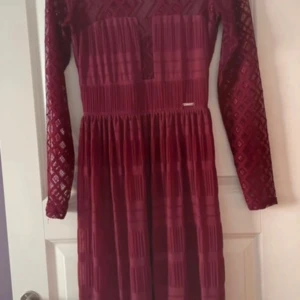 Vinröd långärmad klänning från Guess - Vinröd klänning från Guess med långärmade spetsärmar och geometriskt mönster. Klänningen har rund halsringning och markerad midja, samt dekorativ metallogo framtill. Aldrig använd.