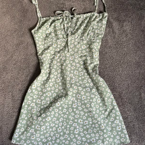 Grön blommig kortklänning med knytband - Söt grön klänning med vita småblommor och smala axelband som knyts upptill. Klänningen har en lätt figurnära passform och är perfekt för varma dagar. Mjuk och tunn känsla i materialet.