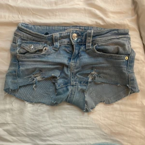 Ljusblå jeansshorts från Gina Tricot - Säljer ett par ljusblå jeansshorts från Gina Tricot med råa kanter och slitna detaljer framtill. Shortsen har klassisk femficksdesign, knapp och dragkedja samt bälteshällor. Perfekta för sommaren!