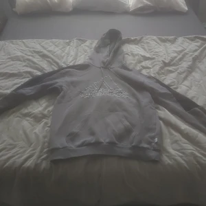 Grå hoodie från Adidas - Säljer en grå hoodie från Adidas med stor logga framtill. Tröjan har huva och en stor magficka. Perfekt för en avslappnad stil och passar bra till vardags. Klassisk modell med långa ärmar och mjukt material.