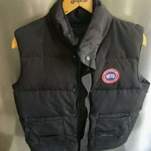 Svart dunväst från Canada Goose - Säljer en svart dunväst från Canada Goose med klassisk logotyp på bröstet. Västen har hög krage, knappar och dragkedja framtill samt två stora fickor. Perfekt för kyliga dagar och riktigt snygg till lager-på-lager-stil.
