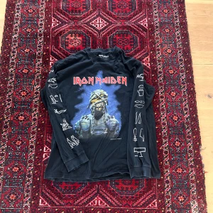 Iron Maiden långärmad t-shirt - Säljer en svart långärmad Iron Maiden t-shirt med stort tryck av bandets ikoniska maskot på framsidan och coola vita tryck längs ärmarna. Perfekt för dig som gillar rock och vill sticka ut med en unik bandtröja.