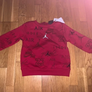 Röd sweatshirt från Jordan med tryck - Cool röd sweatshirt från Jordan med svarta tryck av loggor, text och siffran 23 över hela tröjan. Tröjan har rund hals och långa ärmar. Perfekt för dig som gillar sportig stil och streetwear. Tröja för bebis 