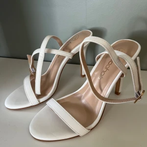 Vita sandaler med klack från Boohoo - Säljer ett par stilrena vita sandaler med smala remmar och öppen tå från Boohoo. Skorna har en elegant klack och justerbart spänne vid ankeln. Perfekta för sommarens alla tillfällen.