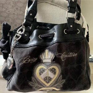 Juicy Couture väska - Juicy Couture axelväska i svart sammet med läderdetaljer och silverfärgade metalldelar. Framsidan har ett broderat emblem med hjärta och krona samt texten Juicy Couture. Väskan har dekorativ nyckelring och J-logga på baksidan.🩷