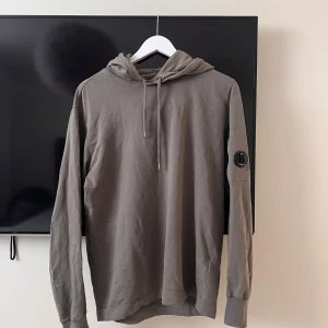 Grå hoodie från C.P. Company - Säljer en grå hoodie från C.P. Company med huva och dragsko. Tröjan har en unik detalj med en rund lins på ena ärmen. Tillverkad i mjuk bomull, perfekt för en avslappnad stil.