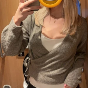 Beige omlottkofta från About You - Mysig beige omlottkofta från About You med knytband i midjan. Koftan har lång ärm och är stickad i ett mjukt material, lite nopprig men inget jag tycker stör! Passar S-L