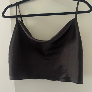 Svart satin croppad linnetopp - Säljer en svart croppad linnetopp i satin med tunna axelband och snygg drapering vid halsen. Perfekt för att lyfta vilken outfit som helst med en stilren och trendig look.