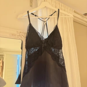 Svart spetslinnetopp från new yorker - Säljer en svart linnetopp från new yorker med tunna axelband och vacker spetsdetalj framtill och på sidorna. Toppen har en liten rosett framtill och är tillverkad i ett mjukt, glansigt material. Perfekt för lager-på-lager eller som statement-piece.