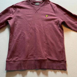 Vinröd sweatshirt från Lyle & Scott - Vinröd sweatshirt från Lyle & Scott med rund halsringning och klassisk gul logga på bröstet. Tröjan har ribbade muddar vid ärmslut och nederkant. Perfekt för en avslappnad stil.