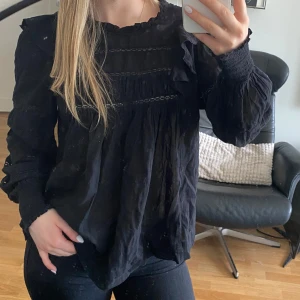 Svart blus med volangdetaljer - Säljer en svart blus med volanger och rynkade ärmar. Blusen har lång ärm och fina spetsdetaljer framtill. Perfekt för dig som gillar en romantisk och trendig stil.