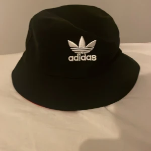 Svart och röd bucket hat från Adidas - Säljer en vändbar bucket hat från Adidas. Ena sidan är svart med vitt broderat Adidas-logo, andra sidan är röd med svart broderat Adidas-logo. Klassisk form och bred skärm, tillverkad i bomull.