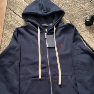 Polo Ralph lauren - Mörkblå hoodie med dragkedja från Polo Ralph Lauren. Tröjan har huva med vita snören, röd broderad logga på bröstet och mjukt foder. Perfekt för en avslappnad stil.