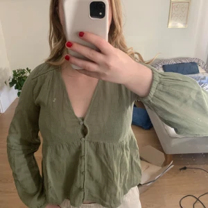 Olivgrön blus/skjorta med knappar - Säljer en olivgrön blus med v-ringning och knappar framtill. Blusen har långa ärmar med lätt puff och är tillverkad i ett tunt, vävt material. Köpt på zalando och aldrig använd med prislapp kvar!