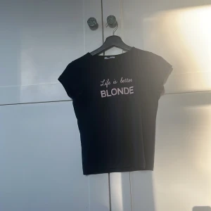 Svart t-shirt med tryck från Subdued - Svart t-shirt från Subdued med texten 'Life is better BLONDE' i rosa framtill. T-shirten har korta ärmar och en klassisk rund halsringning. Perfekt för dig som gillar statement-plagg och enkel stil.