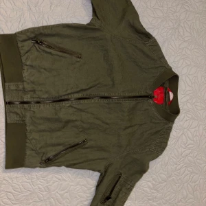 Olivgrön bomberjacka från Zara Man - Säljer en olivgrön bomberjacka från Zara Man. Jackan har dragkedja framtill, två snedställda fickor med dragkedja och en extra ficka på ärmen. Ribbad mudd vid krage, ärmslut och nederkant.