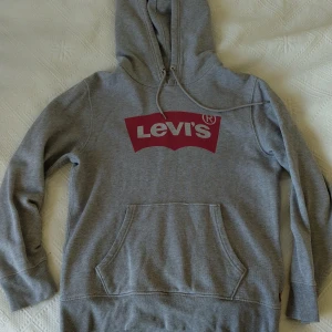Levi's hoodie - Säljer en klassisk grå hoodie från Levi's med röd logga framtill. Tröjan har huva med dragsko och en stor magficka. Perfekt för en avslappnad och stilren look. Storlek xs unisex I mycket gott skick!