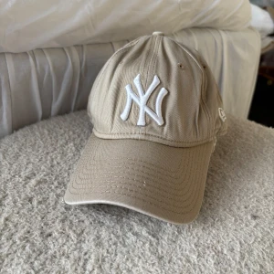 Beige New York Yankees keps från New Era - Snygg beige keps från New Era med New York Yankees-logga broderad i vitt framtill. Justerbar passform med spänne bak och klassisk böjd skärm. Perfekt accessoar för en sportig och avslappnad stil.