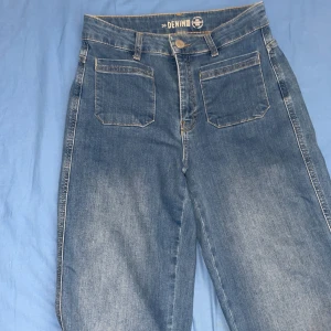 Blå wide leg jeans med hög midja - Säljer ett par blå jeans från Denim 1963 med hög midja och raka, vida ben. Jeansen har klassiska fickor fram och bak samt knapp och dragkedja i gylfen. Perfekta för en avslappnad och trendig look. Jag har även haft på mig den 2-3 gånger. Den är dock ganska gammal, men ser nytt ut.