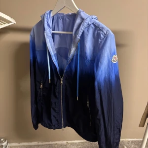 Blå vindjacka från Moncler med huva - Säljer en snygg blå vindjacka från Moncler med gradient från ljusblått till mörkblått. Jackan har huva med dragsko, dragkedja framtill och Moncler-logga på ärmen. Perfekt för dig som vill ha en stilren och lätt jacka. Skriv även om ni har ett prisförslag till mig. Självklart äkta kvitto följer med till den som köper, byten är tillåtna