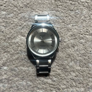 Silverfärgad klocka - Stilren klocka från Regal med rund silverfärgad urtavla och matchande metallarmband. Urtavlan har minimalistisk design med tydliga markeringar och smala visare. Perfekt accessoar för dig som gillar en klassisk look.
