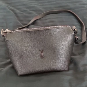 Svart väska från Yves Saint Laurent (fejk)  - Säljer en stilren svart väska från Yves Saint Laurent (fejk) i skinn med silverfärgad dragkedja och YSL-logga framtill. Väskan har en bär rem och ett rymligt huvudfack. Mått: Bred 23 cm. Höjd 17. Djup 9,5. Hör av dig vid frågor 🫶🏽☺️
