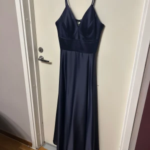 Mörkblå långklänning från dressly  - Elegant mörkblå långklänning med smala axelband och v-ringad front. Klänningen har snörning i ryggen och är tillverkad i ett satin material. Perfekt för bal eller festliga tillfällen. Den är även justerbar i ryggen så den kan funka för lite mindre storlek 