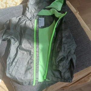 Grå vindjacka med neongröna detaljer från Ellos - Snygg grå vindjacka från Ellos med neongröna dragkedjor och foder. Jackan har huva och resår vid ärmslut samt praktiska fickor framtill. Perfekt för blåsiga dagar och enkel att matcha med andra plagg. Jackan är i storlek 98!