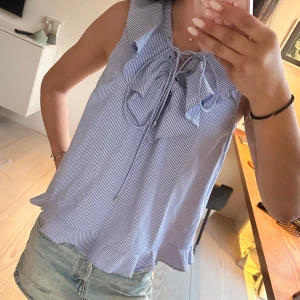 Blåvit randig ärmlös blus med volang och knyt - Säljer en söt blåvit randig blus med volangdetaljer och knyt framtill. Blusen är ärmlös och har en luftig passform, perfekt för varma dagar. Passar lika bra till jeansshorts som till kjol.