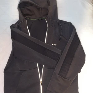 Grå kofta från BOSS - Snygg grå hoodie från BOSS med dragkedja och huva. Jackan har diskret logga på bröstet, ribbade detaljer på ärmarna och fickor framtill. Perfekt för en avslappnad stil.