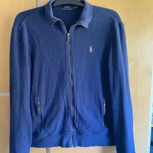 Blå zip-jacka från Polo Ralph Lauren - Säljer en blå zip-jacka från Polo Ralph Lauren med klassisk krage och broderad logga på bröstet. Jackan har två fickor med dragkedja och ribbade muddar. Perfekt för dig som gillar stilren och sportig look.