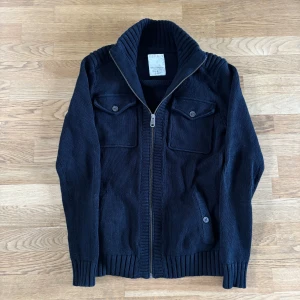 Svart stickad tröja från Esprit - Svart stickad jacka med grov ribbad krage och dragkedja framtill. Två bröstfickor och två sidofickor med knappar. Jackan har lång ärm och ribbade muddar vid ärmslut och nederkant. Perfekt för kyliga dagar.