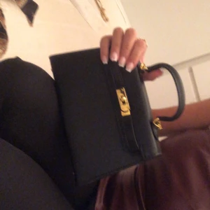 Svart handväska med gulddetaljer - Säljer en stilren svart handväska med guldfärgat spänne och detaljer. Väskan har ett klassiskt och elegant utseende med ett handtag upptill. Perfekt för dig som vill ha en snygg accessoar till din outfit.