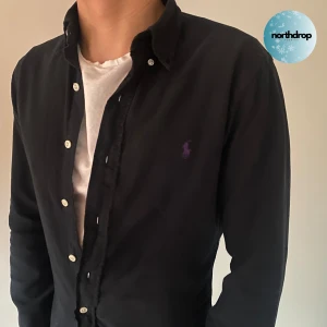 Svart långärmad skjorta från Ralph Lauren - Snygg svart långärmad skjorta från Ralph Lauren med klassisk krage och knappar framtill. Perfekt för en stilren look. Den har en broderad logga i lila på bröstet. Skjortan är i mycket bra skick, men saknar en av knapparna vid ena handleden, dock inget man tänker på och går att knäppa med den andra knappen.